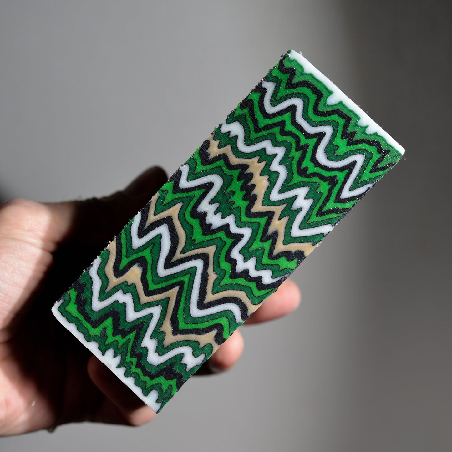 Handmade custom micarta block featuring a sharp zigzag pattern in vibrant green, black, white, and tan colors for knife scales