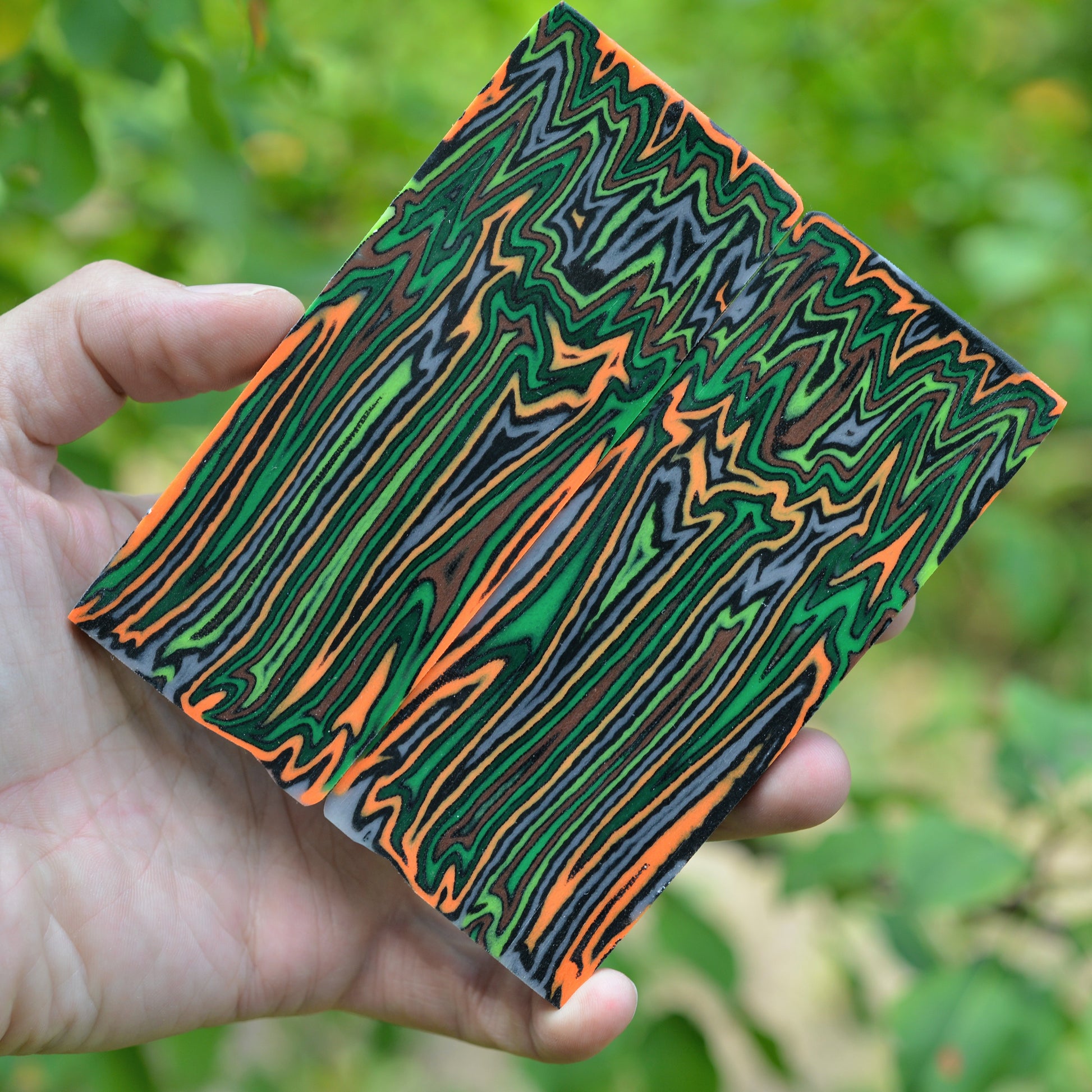 Handmade micarta block with green, orange, black, and gray layered stripes held outdoors.