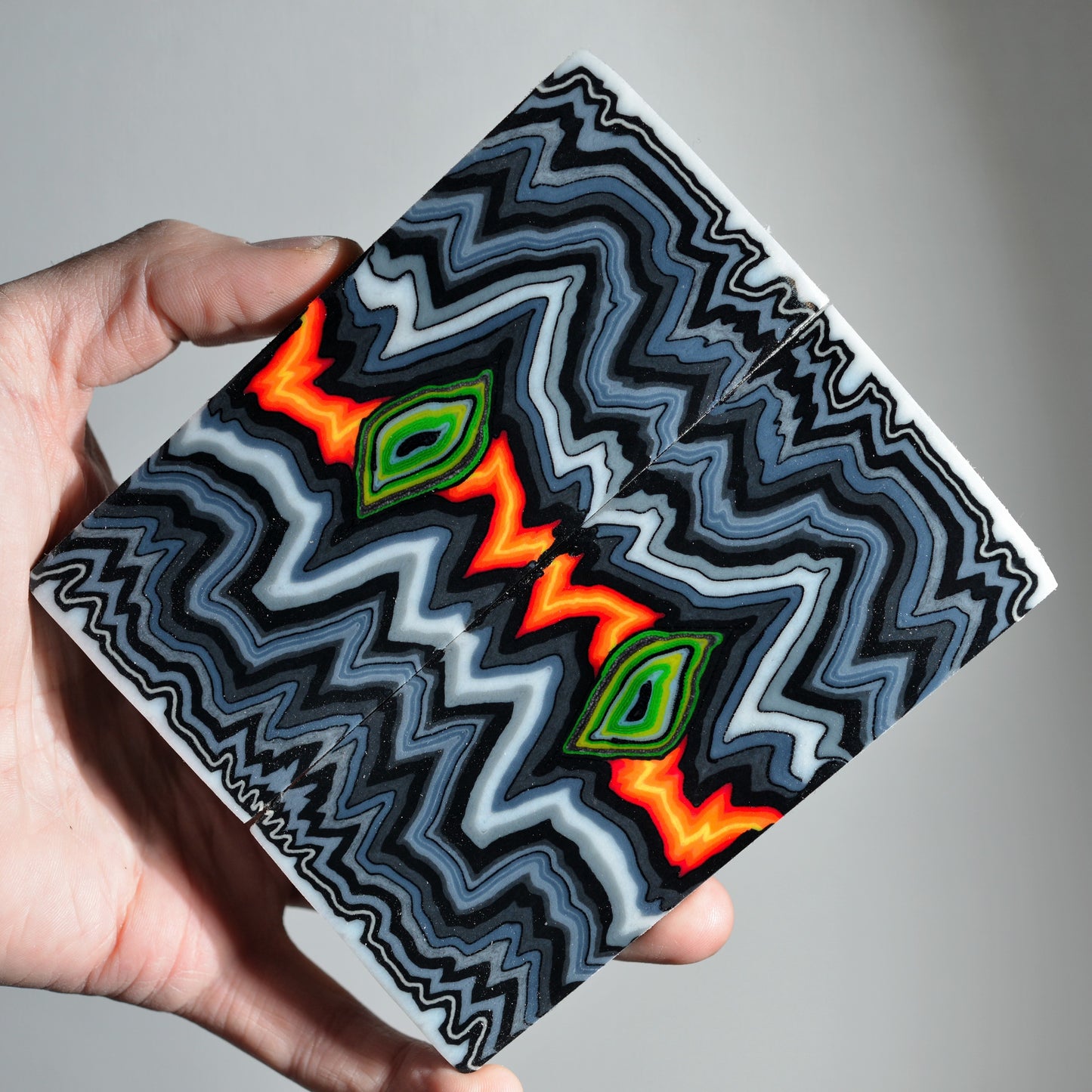 A pair of premium custom micarta knife scales featuring a symmetrical, hypnotic zigzag pattern in black, grey, and white with vibrant neon orange and green "eye" centers.
