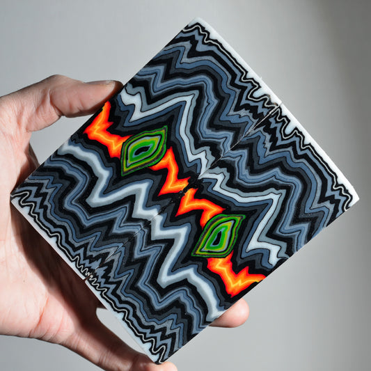 A pair of premium custom micarta knife scales featuring a symmetrical, hypnotic zigzag pattern in black, grey, and white with vibrant neon orange and green "eye" centers.