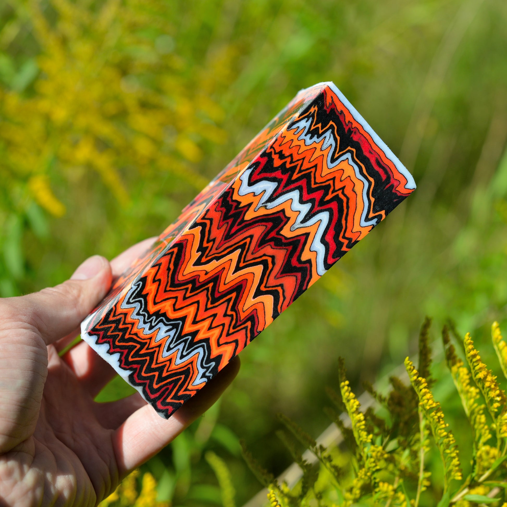 Handmade micarta-style layered composite block pattern in red, yellow, orange and black colors, held in hand for knife handle making